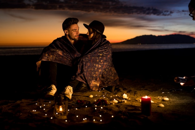 Young couple in blanket sitting on sea shore 