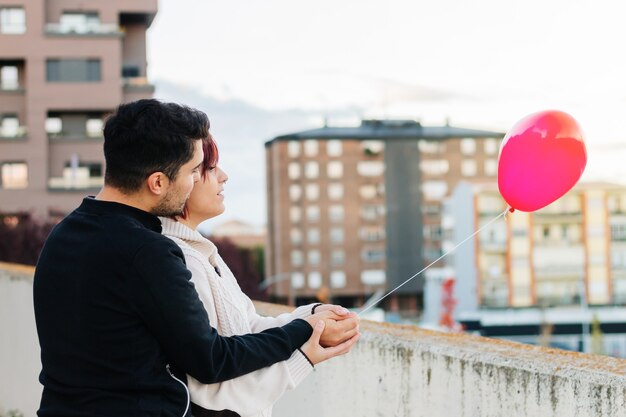 젊은 커플, 소년과 소녀는 등을 돌리고 알아볼 수 없는 상태로 도시를 배경으로 발코니에서 빨간 하트 모양의 풍선을 들고 있습니다. 공간, 사랑 개념, 발렌타인을 복사합니다.
