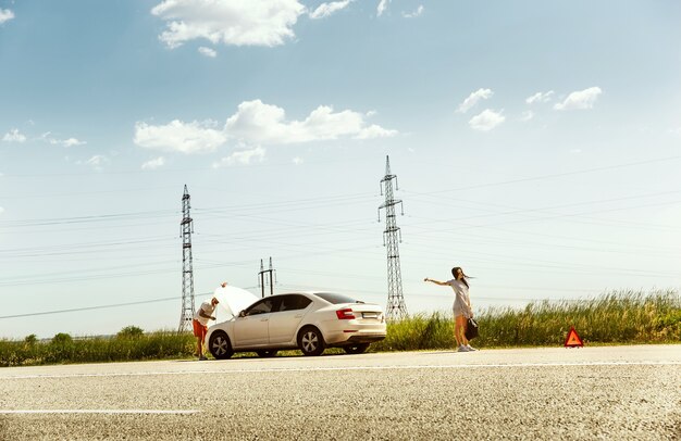 The young couple broke down the car while traveling on the way to rest. They are trying to stop other drivers and ask for help or hitchhike. Relationship, troubles on the road, vacation.