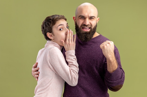 young couple in casual clothes bearded man clenching fist while his girlfriend telling a secret to him celebrating valentines day standing over green wall