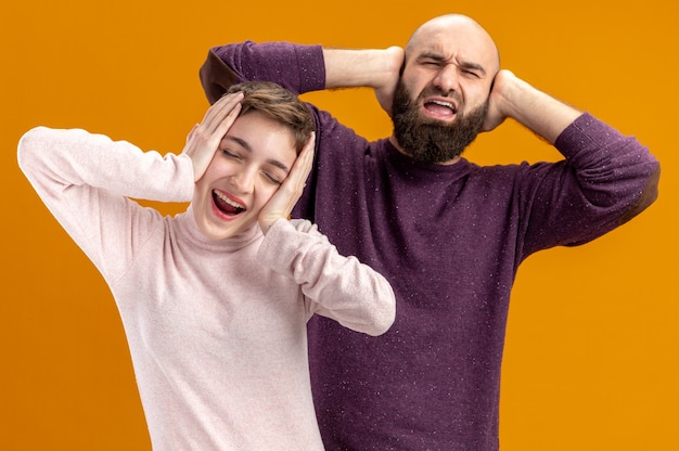 Foto gratuita coppia giovane in abiti casual barbuto uomo e donna con i capelli corti felice ed eccitato tenendo le mani sulle teste il giorno di san valentino concetto in piedi su sfondo arancione