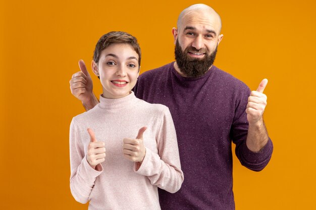 young couple in casual clothes bearded man and woman with short hair looking at camera happy and cheerful smiling showing thumbs up valentines day concept standing over orange wall