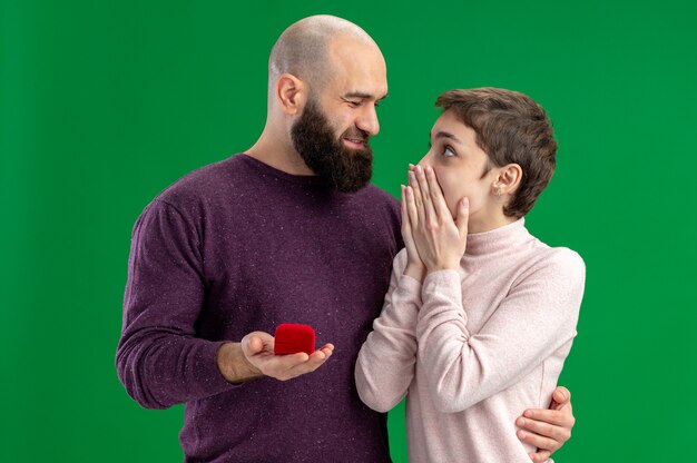 young couple in casual clothes happy and bearded man making proposal with engagement ring in red box to his amazed girlfriend during valentines day standing over green background