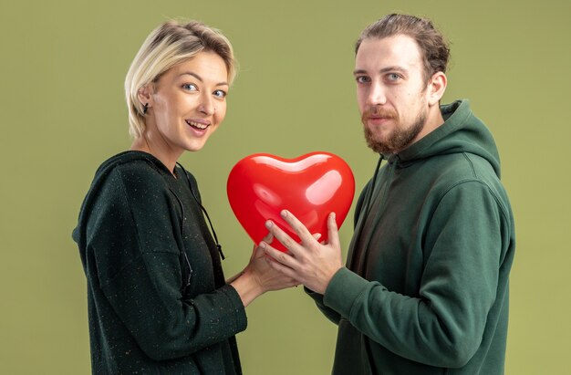 young couple in casual clothes woman and man holding heart shaped balloon together happy in love  smiling cheerfully celebrating valentines day standing over green wall