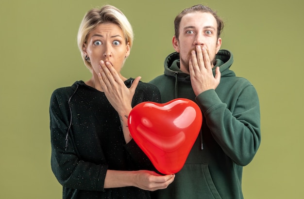 Free photo young couple in casual clothes woman and man with heart shaped balloon looking at camera amazed and surprised covering mouth with hands celebrating valentines day standing over green background