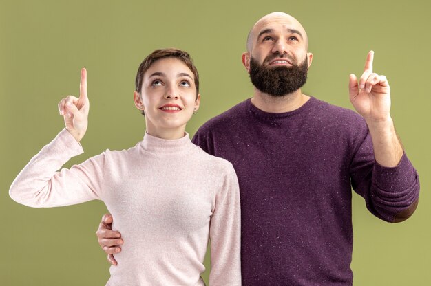 young couple in casual clothes woman with short hair and bearded man looking up showing index fingers happy and positive celebrating valentines day standing over green background