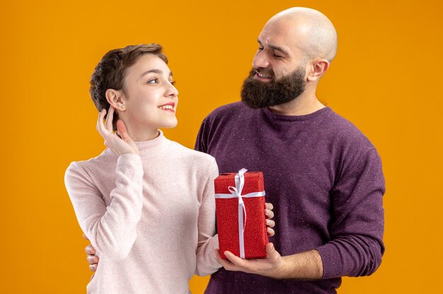 young couple in casual clothes woman with short hair with present and bearded man looking at each other happy in love together celebrating valentines day standing over orange wall