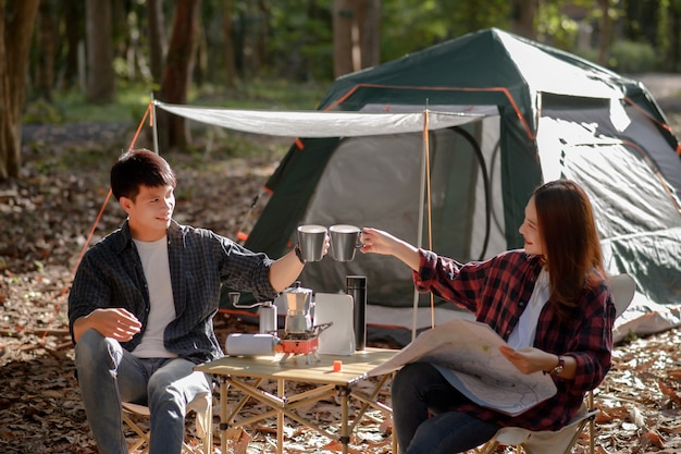 自然公園で朝のキャンプテントの前で朝コーヒーマグを一緒にチリンと鳴らす若いカップル