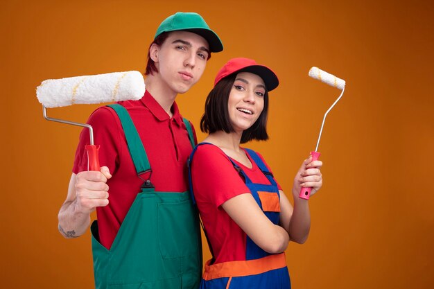 건설 노동자 유니폼을 입은 젊은 부부 자신감 있는 남자와 흥분한 소녀와 주황색 벽에 격리된 카메라를 바라보는 페인트 롤러를 들고 프로필 보기에 서 있는 모자
