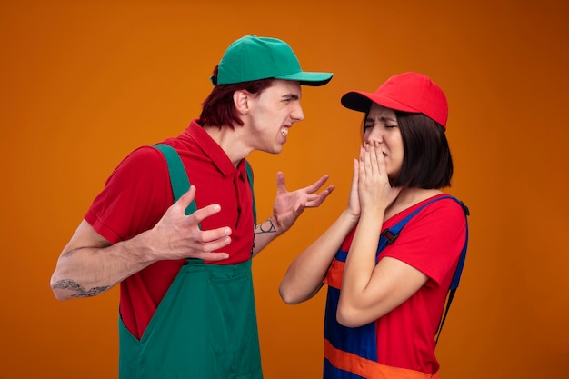 Young couple in construction worker uniform and cap aggressive guy looking at girl spreading hands scared girl keeping hands on mouth with closed eyes 