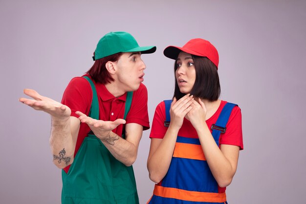 Young couple in construction worker uniform and cap clueless guy showing empty hands looking at girl pointing at side with hands concerned girl keeping hands together looking at side isolated