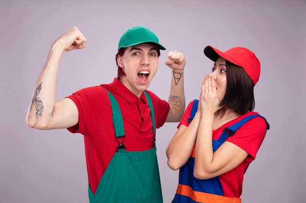 Young couple in construction worker uniform and cap confident guy doing strong gesture concerned girl keeping hands on mouth looking at guy 