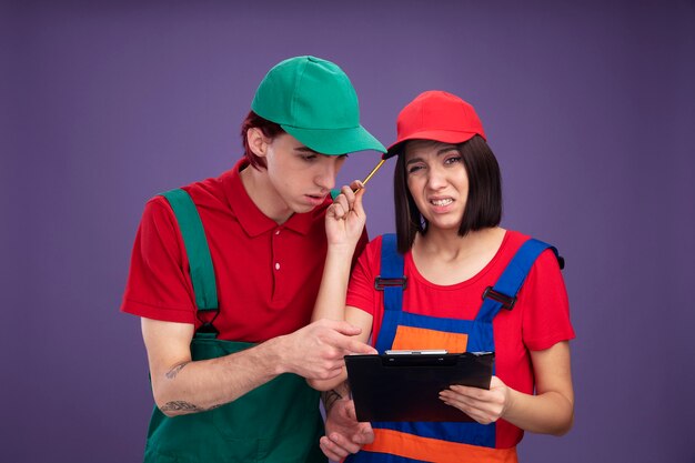 建設労働者の制服とキャップの若いカップルは、鉛筆とクリップボードを持って頭に触れ、鉛筆を集中して男を見て、クリップボードを指差して混乱している
