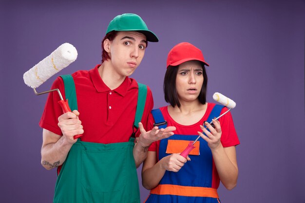 Young couple in construction worker uniform and cap holding paint roller clueless guy looking at camera showing empty hand confused girl looking at her paint roller isolated on purple wall