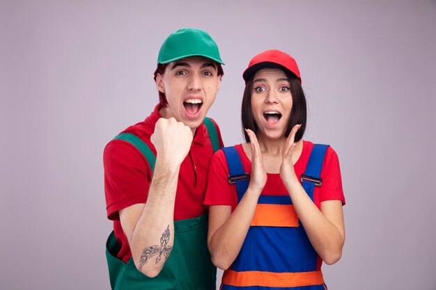 Young couple in construction worker uniform and cap joyful guy doing yes gesture excited girl keeping hands in air both looking at camera isolated on white wall