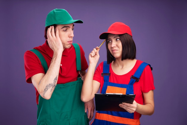 Young couple in construction worker uniform and cap looking at each other unpleased girl holding pencil and clipboard touching head with pencil concerned guy keeping hand on head