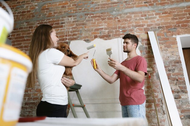 Young couple doing apartment repair together themselves. Married man and woman doing home makeover or renovation. Concept of relations, family, pet, love. Painting the wall, holding the dog.