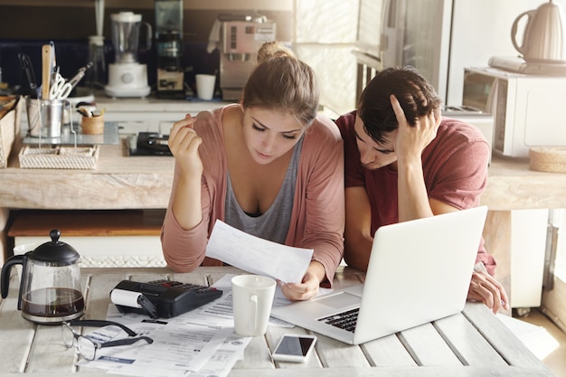 Young couple doing paperwork in kitchen: frustrated woman reading document together with her husband who is holding his head in desperation, sitting at table with laptop