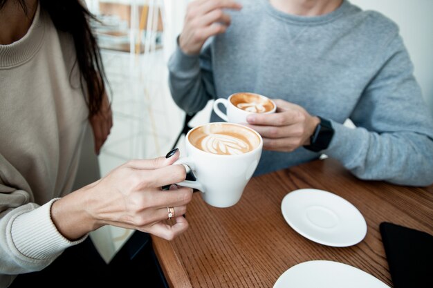 温かい飲み物を飲む若いカップル テーブルの上に暖かいコーヒーのカップを持って カフェに座っている女性と男性を閉じます 居心地の良い喫茶店で週末を一緒に過ごす男女 ロマンチックな日付の概念 プレミアム写真