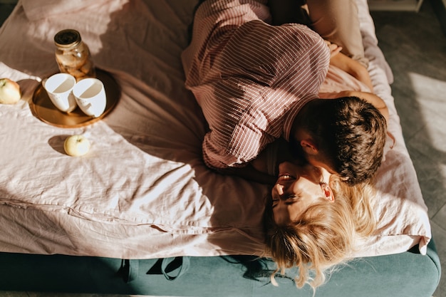 Young couple fooling around in bed during breakfast. Guy in striped shirt hugs laughing girl.