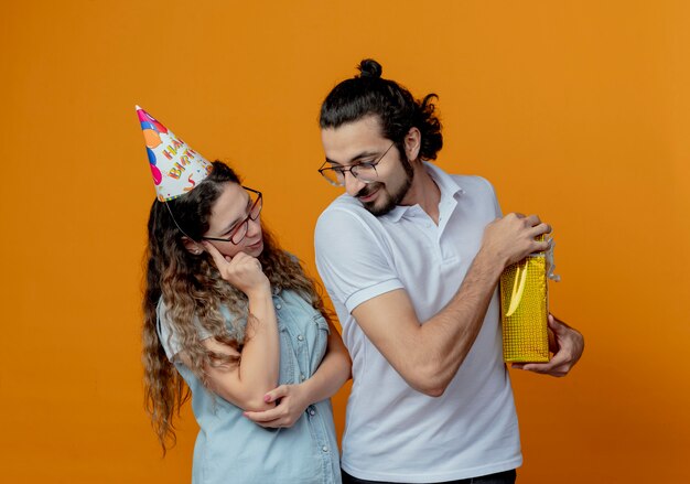 Young couple girl in birthday cap thinking and guy covered gift isolated on orange