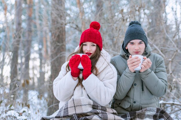 Молодая пара устроила пикник в лесу зимой. влюбленные завернулись в одеяло и пьют чай.