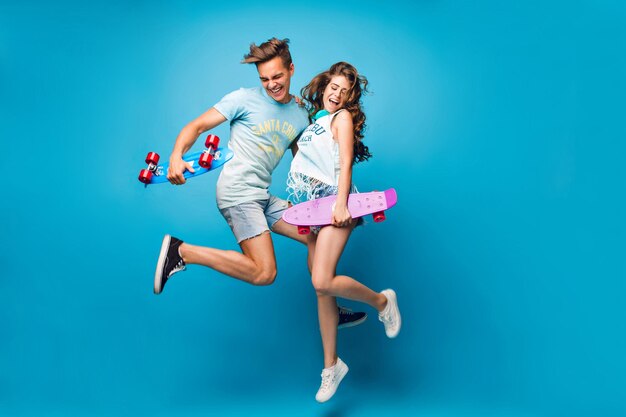 ハンサムな男と長い巻き毛のかわいい女の子の若いカップルは、スタジオで青い背景にジャンプしています。彼らはTシャツを着て、ジーンズを短くし、スケートボードを手に持っています。