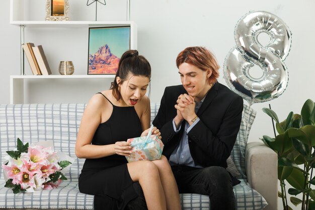 Young couple on happy women day holding and looking at present sitting on sofa in living room