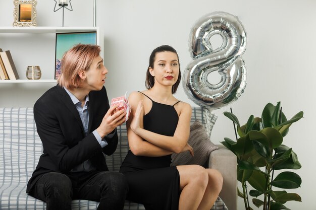 Young couple on happy women day sad guy gives present to strict woman sitting on sofa in living room