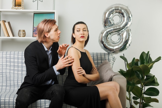 Young couple on happy women day sad guy looking at strict girl sitting on sofa in living room