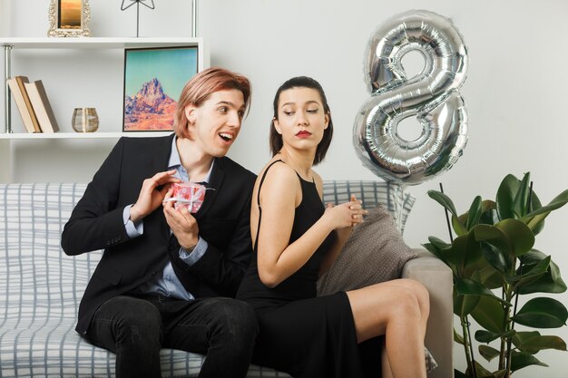 Young couple on happy women day smiling guy gives present to sad girl sitting on sofa in living room