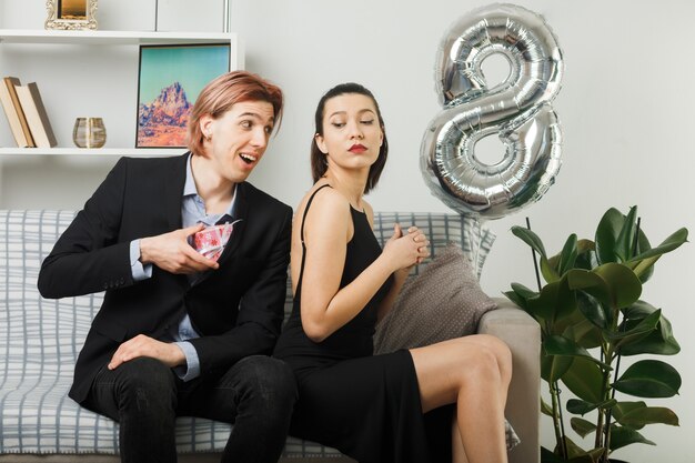 Young couple on happy women day smiling guy gives present to unpleased girl sitting on sofa in living room