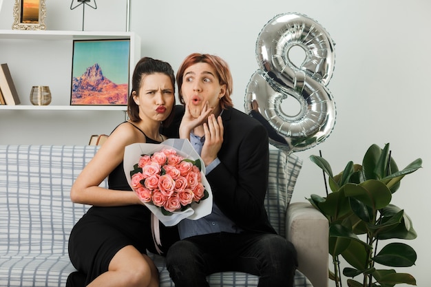 Young couple on happy women day unpleased girl holding bouquet grabbed his chin sitting on sofa in living room