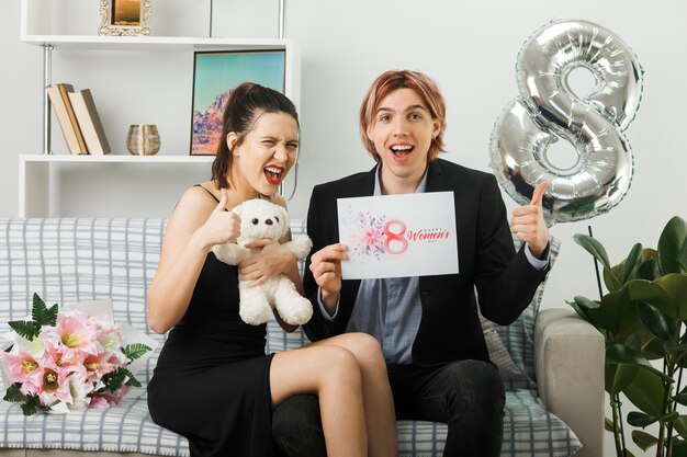 Young couple on happy women day with teddy bear and postcard sitting on sofa in living room