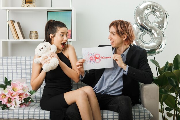 Young couple on happy women day with teddy bear and postcard sitting on sofa in living room