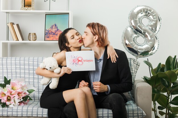 Young couple on happy women day with teddy bear and postcard sitting on sofa in living room