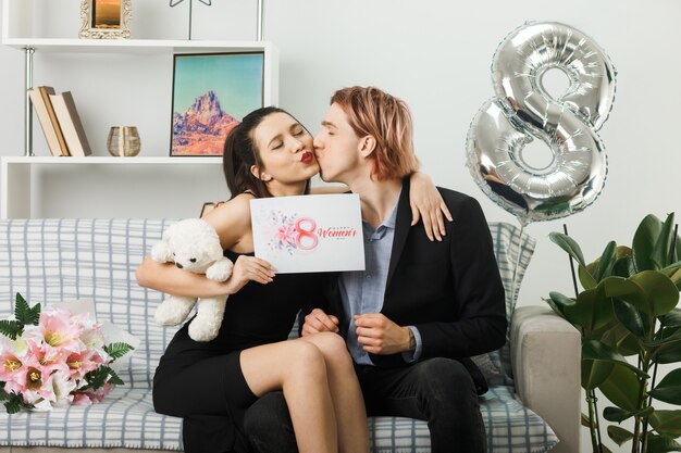 Молодая пара в счастливый женский день с плюшевым мишкой и открыткой, сидя на диване в гостиной