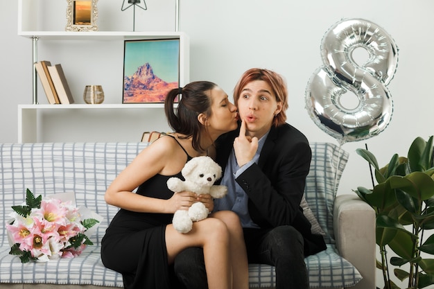 Young couple on happy women day with teddy bear woman kissing his cheek sitting on sofa in living room