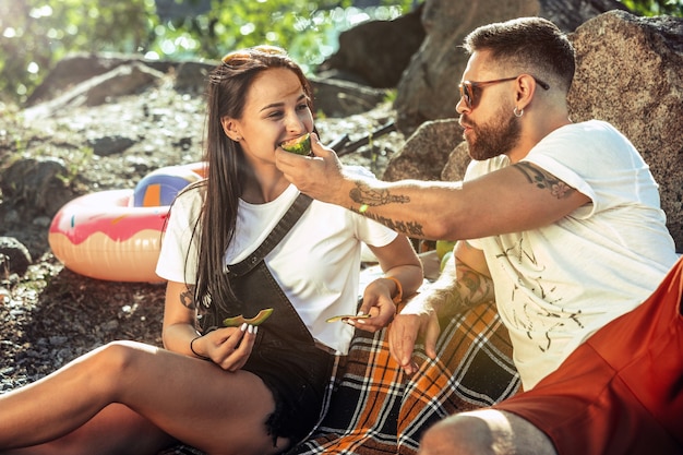 晴れた日に川沿いでピクニックをしている若いカップル。一緒に自然に時間を費やす女性と男性。楽しんで、食べて、遊んで、笑って。関係、愛、夏、週末の概念。