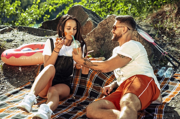 Young couple having picnic at riverside in sunny day. Woman and man spending time on the nature together. Having fun, eating, playing and laughting. Concept of relationship, love, summer, weekend.