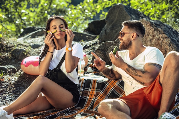 Young couple having picnic at riverside in sunny day. Woman and man spending time on the nature together. Having fun, eating, playing and laughting. Concept of relationship, love, summer, weekend.