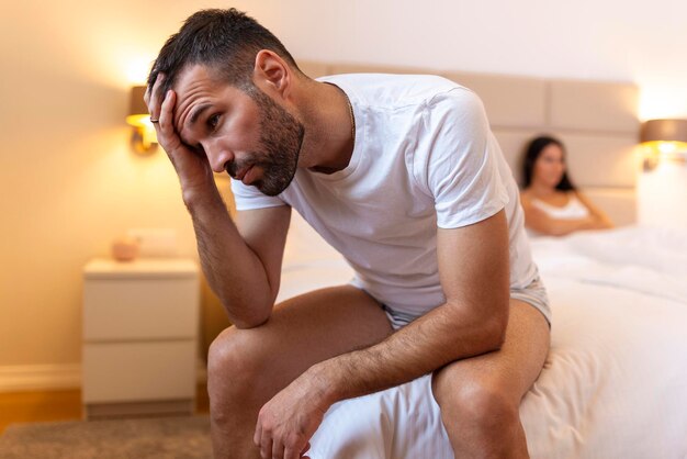 問題を抱えている若いカップル男はベッドに座って、バックグラウンドで彼のガールフレンドを悲しげに見つめていますセックスに問題を抱えている若いカップルを動揺させます