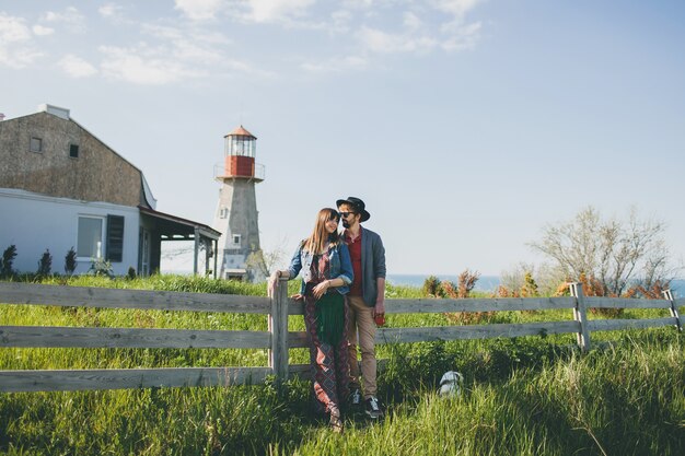 Young couple hipster indie style in love walking in countryside, holding hands, lighthouse on background, warm summer day, sunny, bohemian outfit, hat