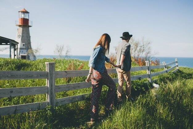 Young couple hipster indie style in love walking in countryside, holding hands, lighthouse on background, warm summer day, sunny, bohemian outfit, hat