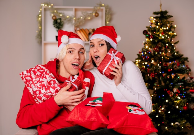 Young couple at home at christmas time wearing santa hat sitting on armchair holding christmas gift sacks and packages impressed guy and surprised girl both looking at camera in living room