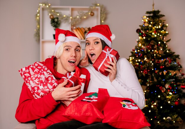 Молодая пара дома на рождество в шляпе санта-клауса, сидя на кресле с рождественскими подарочными мешками и пакетами, впечатлила парня и удивленную девушку, смотрящие в камеру в гостиной
