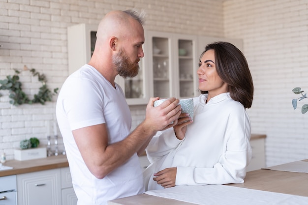 Молодая пара муж с женой дома на кухне, счастливая улыбка смеются, пьют кофе по утрам