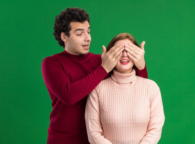 Young couple impressed man standing behind excited woman covering her eyes with hands looking at her isolated on green wall