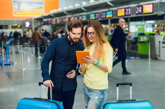 Young couple is standing between two suitcases in airport. She has long hair, glasses, sweater, jeans. He wears beard, black shirt with pants. They are reading on tablet.