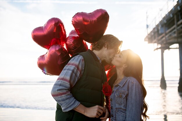 Молодая пара целуется на пляже на закате, держа в руках воздушные шары в форме сердца
